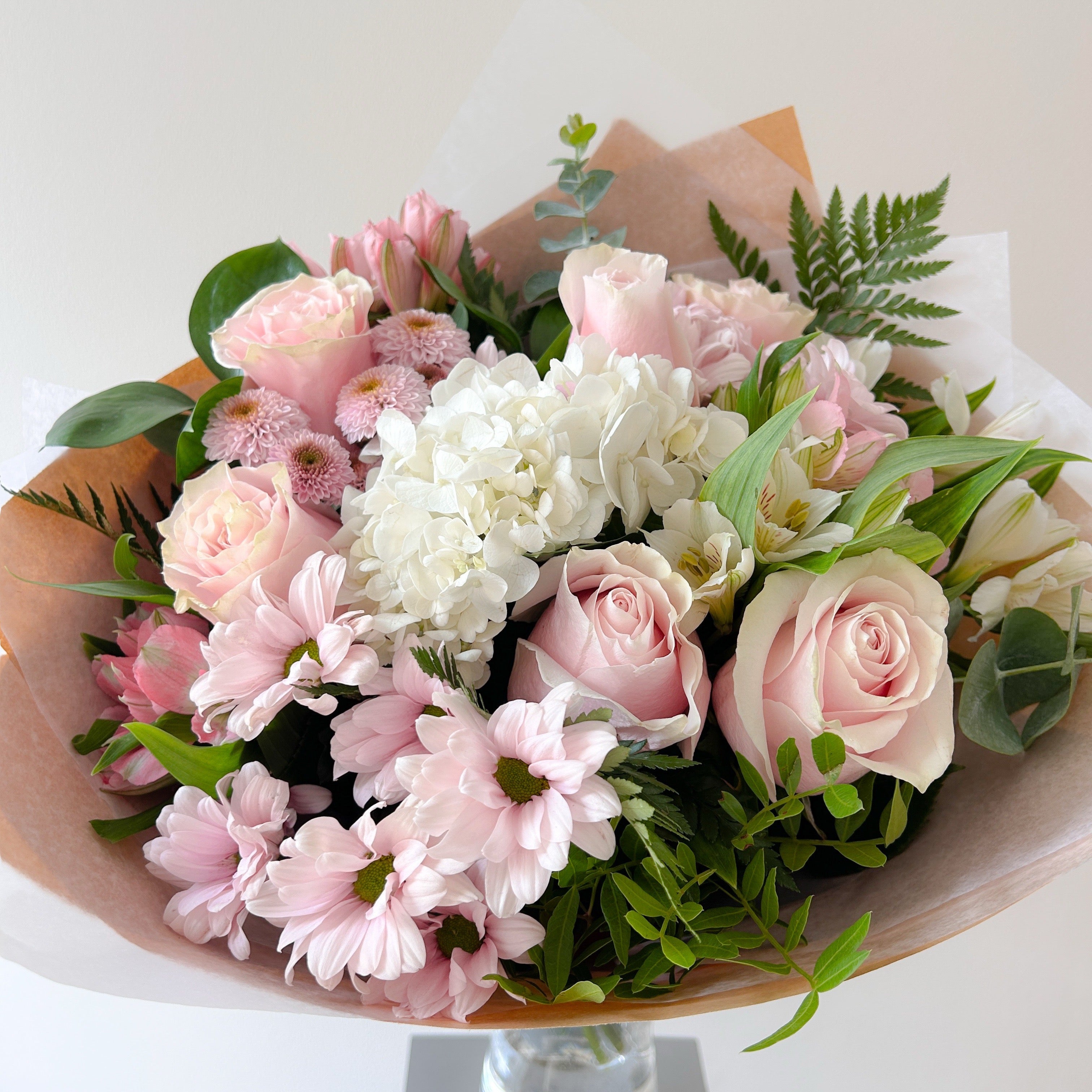 A bouquet of pink and white flowers wrapped in brown paper, featuring light pink roses, white hydrangeas, pink chrysanthemums, and green foliage.