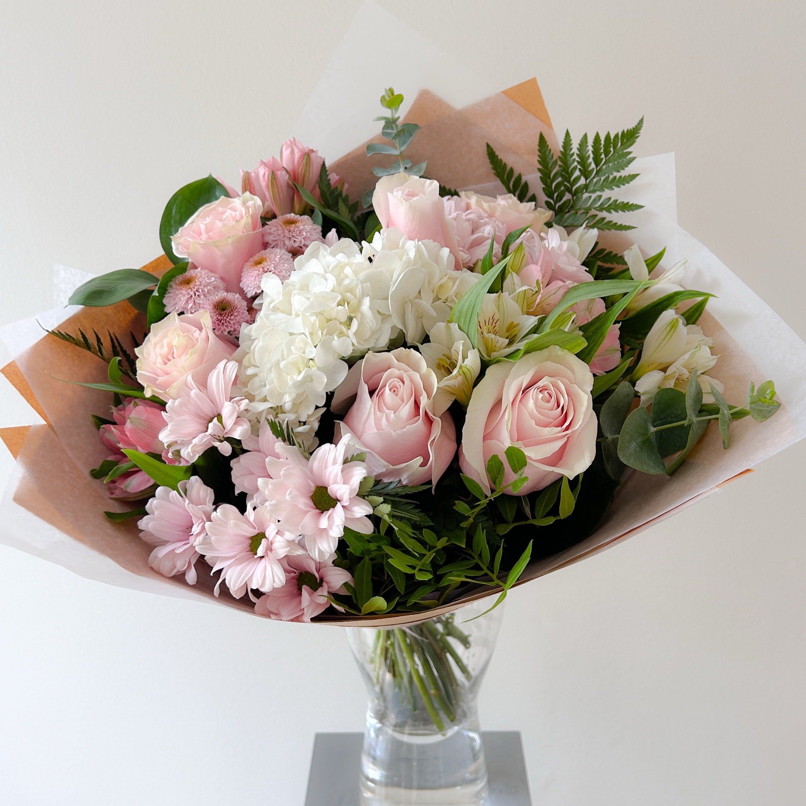 A vibrant floral bouquet featuring pink and white flowers, including roses, and daisies, arranged with green foliage and wrapped in brown and white paper, presented in a clear glass vase on a neutral light background.